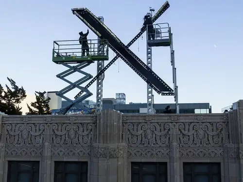 Two workers putting up a large X logo that looks like it was made out of steel beams on top of the Twitter headquarters building.
